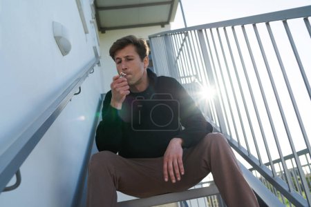 Foto de Hombre joven adulto fumando cigarrillo RYO o marihuana en la escalera junto a su casa - Imagen libre de derechos