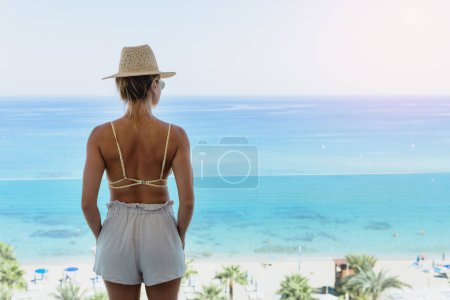 Téléchargez les photos : Jeune femme en bikini et chapeau de paille profiter de la vue depuis le balcon de l'hôtel ou de l'appartement en bord de mer - en image libre de droit