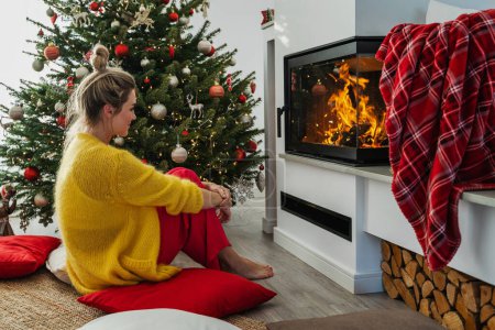 Photo for Young woman sits beside a glowing fireplace in a cozy living room adorned with a Christmas tree and festive decorations. - Royalty Free Image