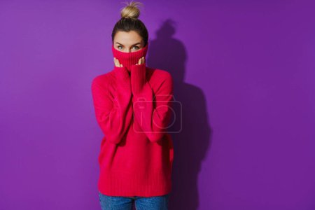 Foto de Retrato de mujer joven está escondiendo su cara dentro de un suéter de cuello de polo cálido y acogedor sobre fondo púrpura - Imagen libre de derechos