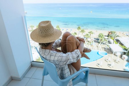 Photo for Young woman relaxing in chair on balcony of beachfront hotel or apartment. - Royalty Free Image