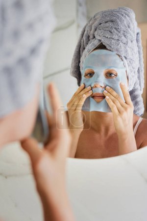 Téléchargez les photos : Belle femme dans la salle de bain avec masque de feuille appliquée sur son visage regardant dans le miroir - en image libre de droit