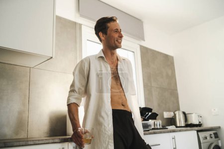 Foto de Retrato de atractivo vago macho bebiendo whisky en su cocina - Imagen libre de derechos