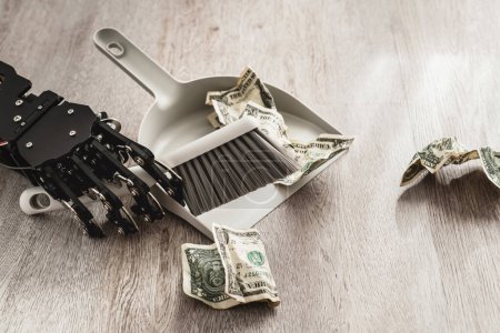 Foto de Mano robótica blandiendo un cubo de basura y un cepillo, barriendo meticulosamente billetes de un dólar. - Imagen libre de derechos