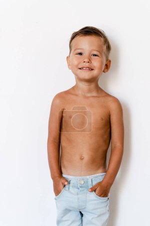 Téléchargez les photos : Portrait de heureux petit garçon joyeux portant un jean posant contre un mur blanc - en image libre de droit