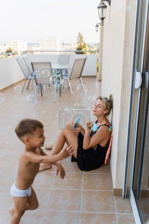 Foto de Alegre madre y su pequeño hijo creando burbujas de jabón en un balcón durante el cálido día de verano. - Imagen libre de derechos