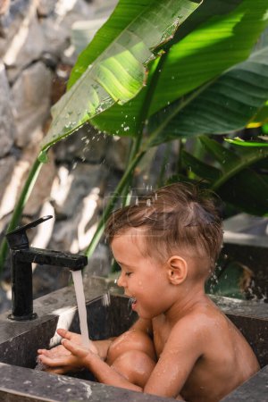 Téléchargez les photos : Adorable tout-petit garçon jouant et se baignant dans un évier en pierre à l'extérieur. - en image libre de droit
