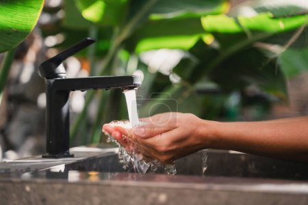 Téléchargez les photos : Femme se lavant les mains avec de l'eau du robinet au lavabo en pierre dans la salle de bain extérieure avec beaucoup de feuilles tropicales - en image libre de droit