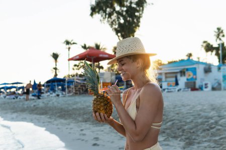 Foto de Joven mujer feliz disfrutando de cóctel en piña durante el atardecer en la playa. - Imagen libre de derechos