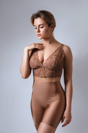 Photo for Portrait of young attractive woman wearing brown lace bra and high waist shaping briefs against gray background. - Royalty Free Image