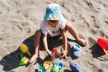 Téléchargez les photos : Mignon petit bambin jouer avec ses jouets en plastique dans le sable sur la plage. - en image libre de droit