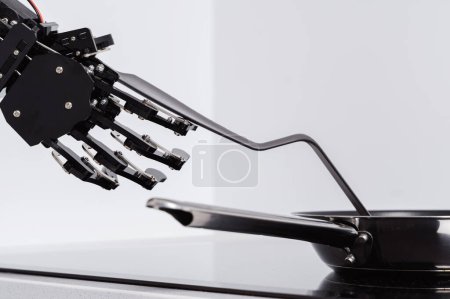 Foto de Auténtica mano robot y sartén en estufa eléctrica. Concepto de automatización robótica de procesos. - Imagen libre de derechos