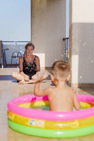 Foto de Madre observa los momentos lúdicos de su hijo pequeño en una piscina inflable en el balcón. - Imagen libre de derechos
