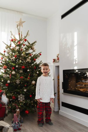Téléchargez les photos : Mignon petit garçon se tient à côté d'une cheminée dans un salon confortable orné d'un arbre de Noël et de décorations festives. - en image libre de droit