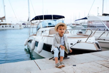 Photo for Cute stylish boy sitting on the pier near yacht in marina. - Royalty Free Image