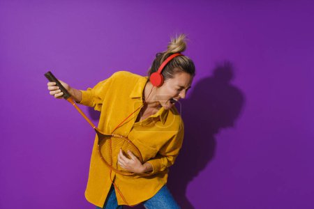 Photo for Cheerful girl is jamming out in her yellow shirt while wearing red headphones. She's strumming away on her racket like it's a guitar, against a vibrant purple background. - Royalty Free Image