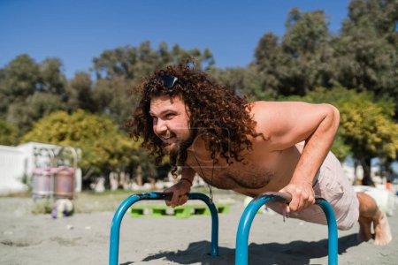 Photo for Curly and bearded man working out on parallel bars during his beach training - Royalty Free Image