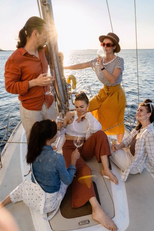 Foto de Grupo de amigos felices bebiendo vino y relajándose en el velero durante la navegación en el mar. - Imagen libre de derechos