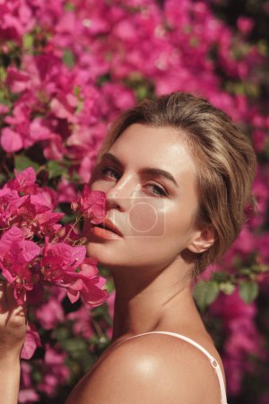 Photo for Beautiful blonde woman with smooth skin and natural makeup, as she poses against a backdrop of a bush with delicate pink flowers. - Royalty Free Image