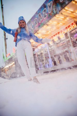 Photo for Cheerful and stylish woman, dressed in warm clothes, is having fun in a snowy winter amusement park. - Royalty Free Image