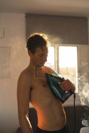 Photo for Portrait of handsome young man ironing his chest with clothes iron - Royalty Free Image