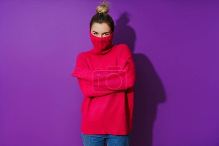 Téléchargez les photos : Portrait de jeune femme cache son visage à l'intérieur d'un pull col polo chaud et confortable sur fond violet - en image libre de droit