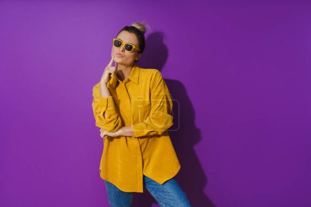 Téléchargez les photos : Portrait de jeune fille portant une chemise jaune et des lunettes de soleil montrant une expression dédaigneuse sur fond violet - en image libre de droit