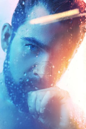 Téléchargez les photos : Portrait de beau jeune homme capturé à travers le verre mouillé dans la lumière bleue et orange. - en image libre de droit