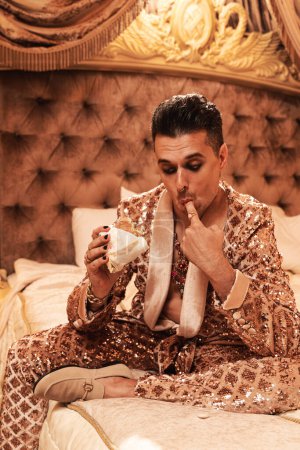 Téléchargez les photos : Homme riche excentrique bizarre vêtu d'un costume doré chatoyant mangeant un hamburger dans une suite royale et luxueuse. - en image libre de droit