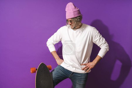 Foto de Retrato de hombre alegre de mediana edad con longboard con sudadera blanca sobre fondo púrpura - Imagen libre de derechos