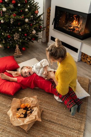Foto de Mujer joven y su pequeño hijo se divierten junto a una chimenea en una acogedora sala de estar adornada con un árbol de Navidad y decoraciones festivas. - Imagen libre de derechos