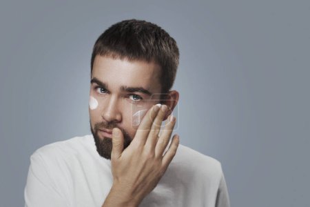 Foto de Hombre joven está aplicando crema hidratante en su cara sobre fondo gris - Imagen libre de derechos