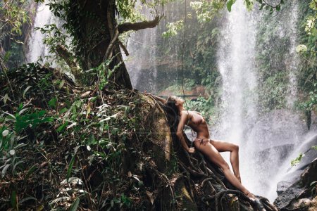 Photo for Wet gorgeous woman posing on the roots tree in the jungle beside mighty Phnom Kulen waterfall in Cambodia - Royalty Free Image