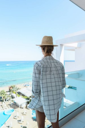 Foto de Mujer joven con sombrero de paja disfrutar de la vista desde el balcón del hotel o apartamento frente al mar - Imagen libre de derechos