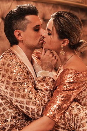 Photo for Wealthy young couple is dressed in radiant golden outfits adorned with sequin embroidery, situated in a luxurious suite. - Royalty Free Image