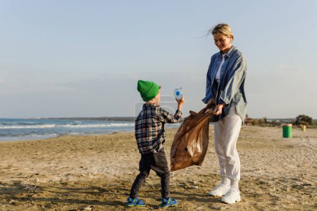 Téléchargez les photos : La famille caucasienne collecte des déchets plastiques sur la plage. Mère enseigne à son fils à garder la nature propre. - en image libre de droit