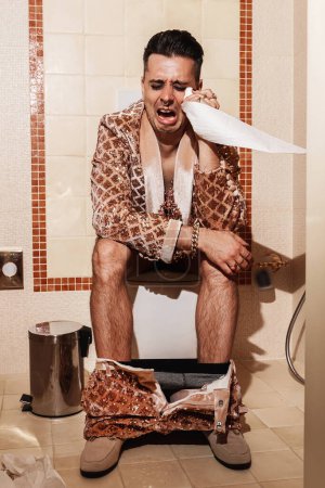 Photo for Rich man, dressed in a radiant golden suit, sits on a toilet and cries. - Royalty Free Image