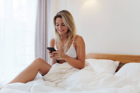 Téléchargez les photos : Jeune femme blonde naviguant sur smartphone au lit pendant les heures du matin. - en image libre de droit
