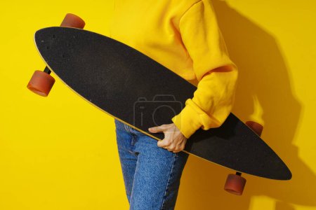 Téléchargez les photos : Femme portant sweat-shirt jaune et jeans tient longboard sur fond jaune - en image libre de droit
