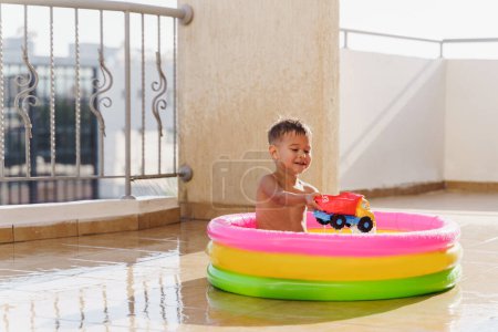 Téléchargez les photos : Tout-petit garçon joue joyeusement dans une piscine ronde gonflable sur le balcon pendant une chaude journée d'été. - en image libre de droit