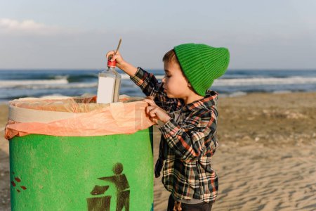 Téléchargez les photos : Mignon garçon jeter bouteille en verre dans la poubelle verte sur la plage pour préserver la propreté. - en image libre de droit