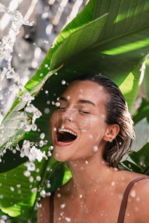 Téléchargez les photos : Jeune femme joyeuse avec des feuilles de banane tropicales et des éclaboussures d'eau - en image libre de droit