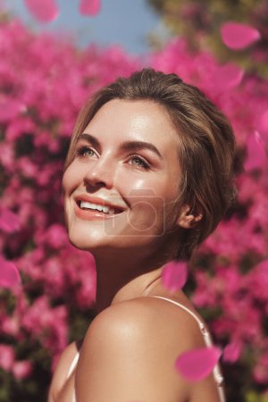Photo for Beautiful blonde woman with smooth skin and natural makeup, as she poses against a backdrop of a bush with delicate pink flowers. - Royalty Free Image