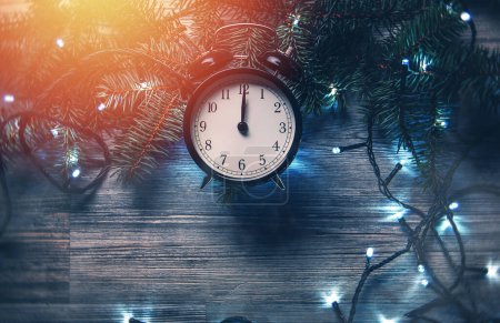Photo for Alarm clock nearing midnight, surrounded by radiant Christmas lights and fresh spruce twigs  on a rustic wooden surface. - Royalty Free Image