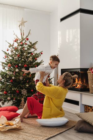 Téléchargez les photos : Jeune femme et son petit fils s'amusent à côté d'une cheminée lumineuse dans un salon confortable orné d'un arbre de Noël et de décorations festives. - en image libre de droit