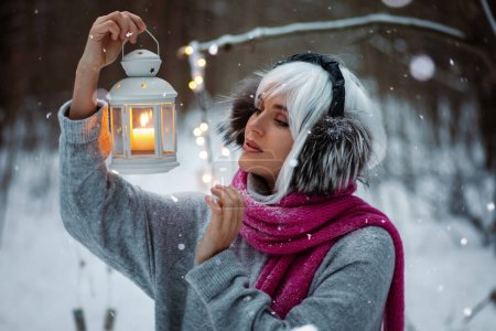 Photo for Woman, adorned in a white wig, thermal earmuffs, cozy sweater, and pink scarf. She holds  a glowing candle lantern. - Royalty Free Image