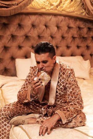 Photo for Freaky eccentric  rich man dressed in a shimmering golden suit eating burger in a royal, luxurious suite. - Royalty Free Image