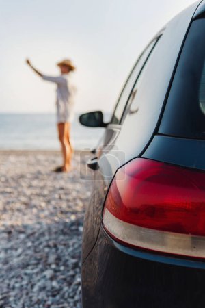 Téléchargez les photos : Femme voyageur se tient à côté de sa voiture sur une plage de galets et capturer la beauté scénique avec son smartphone - en image libre de droit