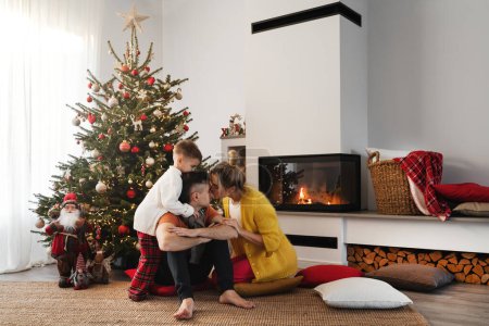 Foto de Joven familia feliz sentarse junto a una chimenea en una acogedora sala de estar, adornada con un árbol de Navidad y decoraciones festivas. - Imagen libre de derechos