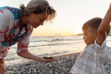 Téléchargez les photos : Mère et son mignon petit fils ramassant des cailloux sur la plage de galets. - en image libre de droit